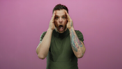 Young man with tattooed arms covers his face with hands expressing sadness against an isolated pink...