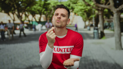 Fototapeta premium Young man with a volunteer shirt gestures for money in a sunny park, reflecting a lighthearted scene outdoors with trees in the background.