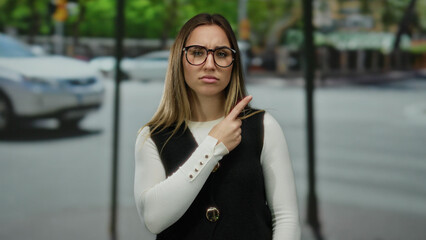Woman pointing with both hands while standing on a busy street surrounded by moving cars and green trees outdoors during daytime.