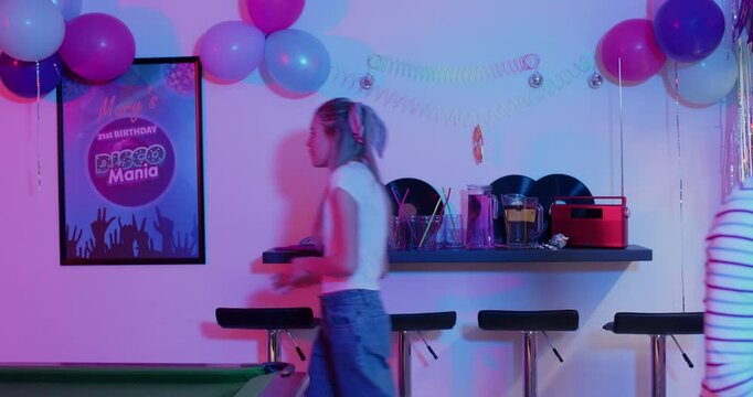 Diverse female friends preparing disco party room, dancing, carrying tray of treats, hugging