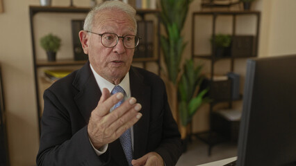 Senior man in suit having a video call in a modern office, surrounded by plants and shelves, expressing animatedly at his computer screen.
