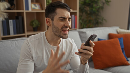 Young hispanic man shouting at phone in modern living room; expressing frustration while holding...
