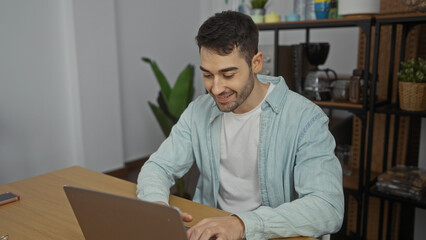 Young hispanic man smiling while working indoors on laptop in office room with plants and wooden furniture, showcasing a productive and modern workplace setting