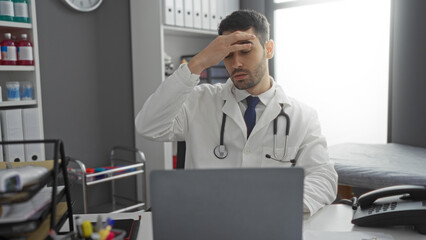 Young male doctor in clinic looks stressed sitting at a desk with computer and stethoscope, appearing fatigued, surrounded by medical supplies and a phone indoors.