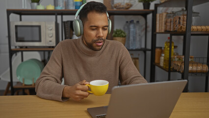Young man with headphones sipping coffee while working on a laptop in an office setting surrounded by kitchenware and modern decor