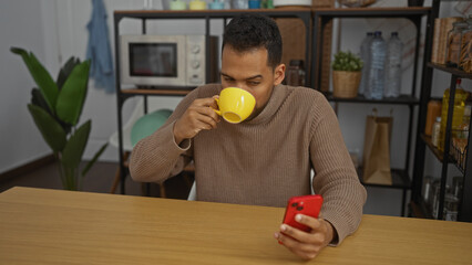Young man sips coffee while texting on smartphone in modern office kitchen with plants and shelves, portraying a casual workplace environment.