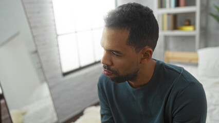 Obraz premium Young hispanic man sitting in a modern bedroom with soft lighting and thoughtful expression, showcasing interior design elements and personal reflection in a domestic setting