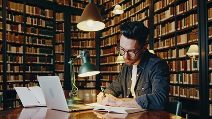 Young college student is taking notes from a textbook while preparing for exams and working on a research paper using his laptop in a traditional old university library - Powered by Adobe