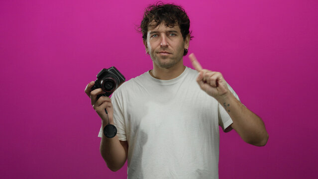 Hispanic man holding camera stands confidently against vibrant pink background, showcasing his photography skills with a hint of artistic flair and an engaging visual presence.