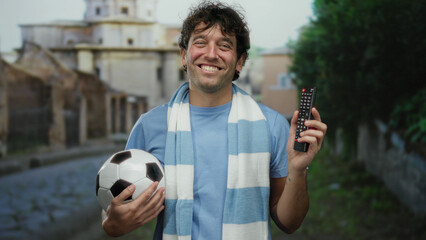 Hispanic man smiling outdoors holding a football and a remote on a street showcasing sports enthusiasm and casual style.