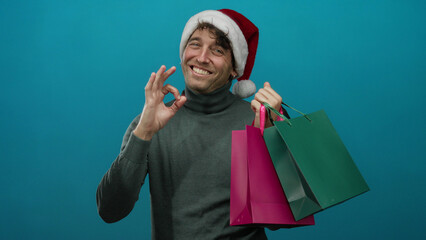Hispanic man in a christmas hat smiles holding shopping bags against a blue background, conveying...