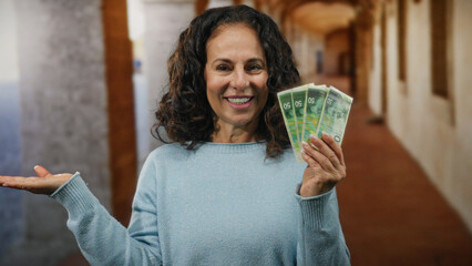 Middle-aged woman in a blue sweater outdoors at a university, holds israeli shekels, smiling with...