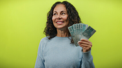 Middle-aged woman holding brazilian real banknotes while smiling against an isolated yellow...