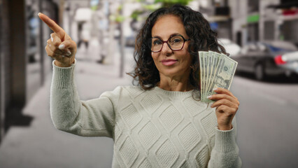 Middle-aged woman holding american dollars outdoors on a city street, confidently pointing in a...