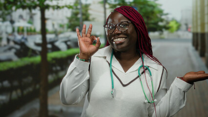 Woman smiling with braided hair and glasses in an outdoor city street, wearing a stethoscope and pointing to her palm, showcasing a vibrant street background.