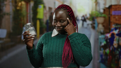 African american woman smiles while holding a jar containing a banknote in a bustling city street...