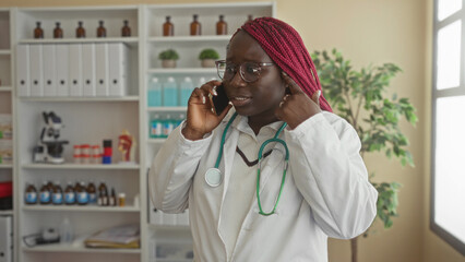 Woman doctor in clinic talking on phone with stethoscope around neck, surrounded by medical equipment, displaying professionalism in healthcare environment.