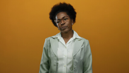 Obraz premium Woman with curly hair and glasses stands confidently against an isolated orange background, wearing a formal blazer, conveying professionalism and seriousness.
