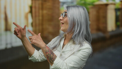 Grey-haired senior woman with tattoos and glasses gestures enthusiastically on an outdoor street...