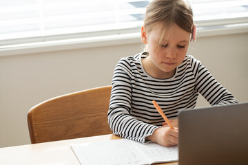 Focused young girl studying with laptop and notepad near window for online education and learning