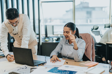 Fototapeta premium Two business employees working together in a modern office setting, discussing ideas and analyzing data on a laptop, fostering teamwork and professional collaboration.