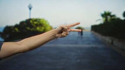 Man showing peace sign with hand in an urban street setting with blurred trees and pathway under a clear sky, evoking a sense of freedom and positivity outdoors.