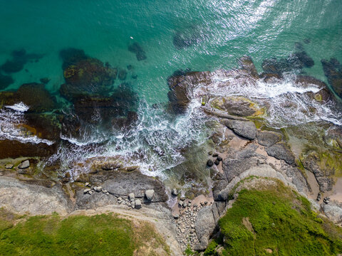 Aerial top-down view of turquoise sea waves crashing over rocky coastline with moss and clear water. Wild nature texture along the Bulgarian coast, dramatic and scenic