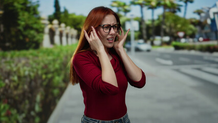 Woman outdoors with glasses and red hair covers ears from noise on city street with green scenery...