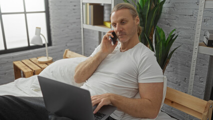 Middle-aged man with blonde hair using laptop and smartphone in cozy bedroom interior by a window...
