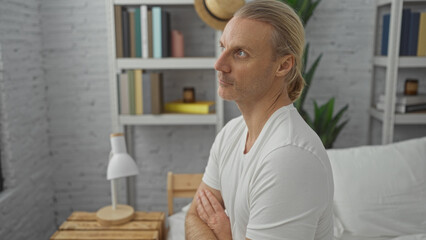 Middle-aged caucasian man with blonde hair sitting on bed in a modern bedroom interior, looking...