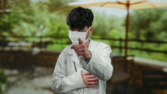 Young man wearing white coat and mask laughing in park under umbrella with arms crossed amidst lush greenery