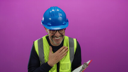 Smiling hispanic mature man in a hard hat and safety vest holding a clipboard stands against an isolated pink background