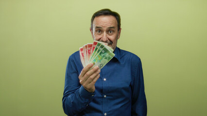 Man holding israeli shekels in hand with a thoughtful look against a yellow background, suggesting...