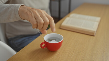 Man making tea in a red mug while reading a book at an office table, showcasing a peaceful moment in an indoor workspace setting.