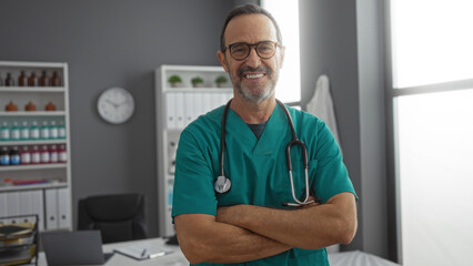 Hispanic male doctor smiling confidently with arms crossed in a well-organized hospital room,...