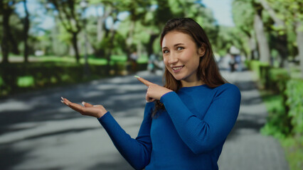 Young woman in blue sweater pointing and giving thumbs up in a sunny park, surrounded by trees and blurred people.