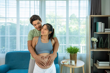 Loving Asian couple man and woman embracing pregnancy with heart hands on belly in cozy home