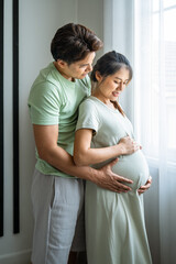 Tender moment of expecting Asian couple man and woman embracing pregnancy near window