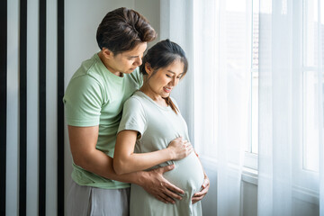 Tender moment of expecting Asian couple man and woman embracing pregnancy near window