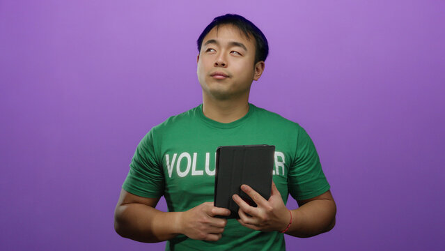 Young man in green volunteer shirt holds tablet against purple background, showcasing an isolated setting emphasizing community involvement and digital engagement.