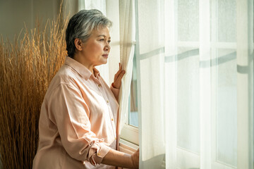 Peaceful Asian senior woman looking out window with morning sunlight and calm expression