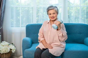 Senior Asian woman exercising at home with dumbbell for healthy aging and strength, looking at camera