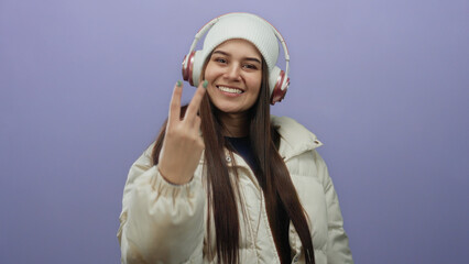 Woman wearing headphones and winter coat celebrates while standing isolated against a bright purple background, expressing joy and energy with a playful gesture.