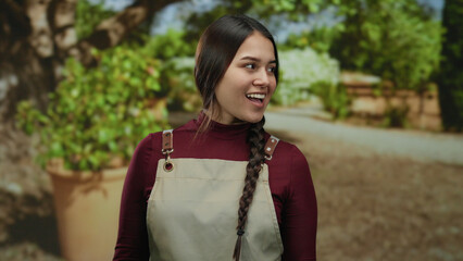 Fototapeta premium Woman smiling in outdoor park setting wearing apron, suggesting her role as waitress, with greenery and natural daylight enhancing cheerful atmosphere.