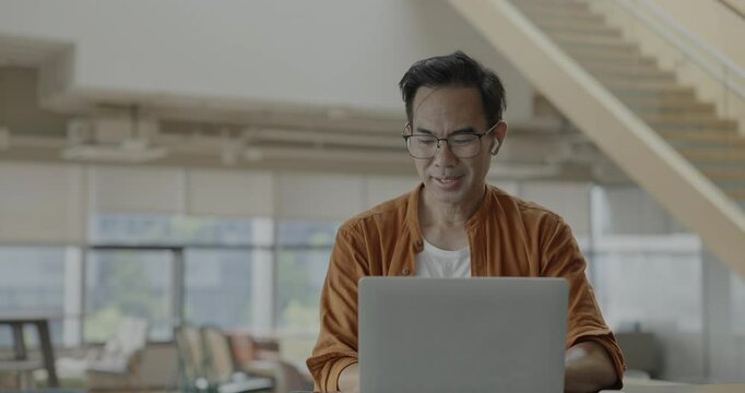 Senior Asian businessman wearing headset celebrates success during online meeting in modern co-working space. Remote work and business communication concept.	