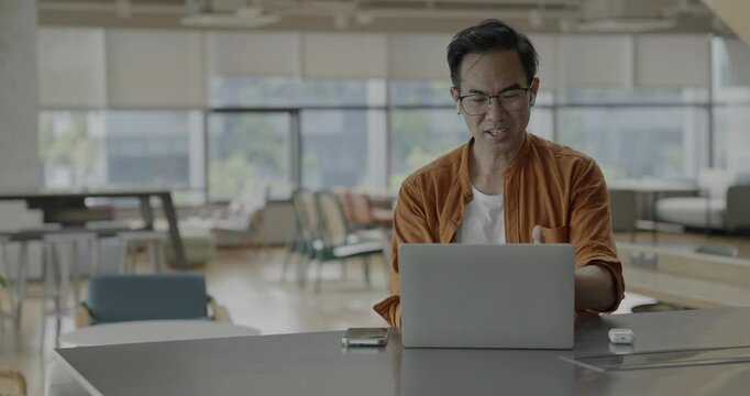 Senior Asian businessman wearing headset celebrates success during online meeting in modern co-working space. Remote work and business communication concept.	
