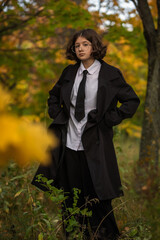 Portrait of a teenage girl wearing glasses and a black coat, standing in an autumn park with colorful fall foliage in background