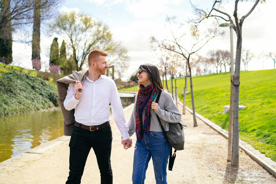 Happy young couple holding hands and walking in a park by the river enjoying a sunny spring day - Powered by Adobe