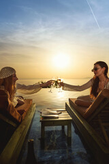 A brunette and blonde in their 20s clink champagne glasses on sunbeds during a picturesque poolside sunset