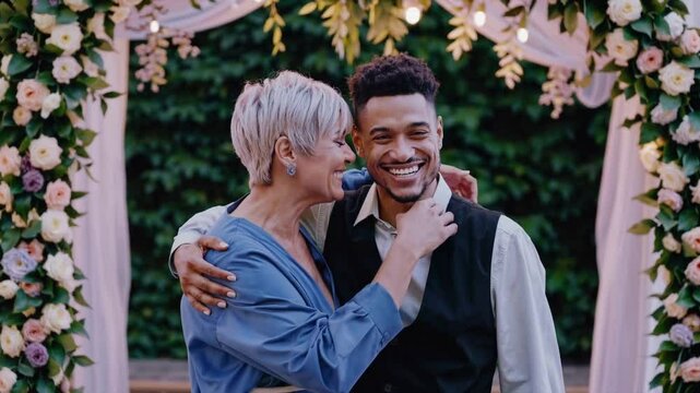 A joyful couple embraces under a floral arch. The woman has short gray hair and wears a blue dress. The man has curly hair and is dressed in a black vest and white shirt.